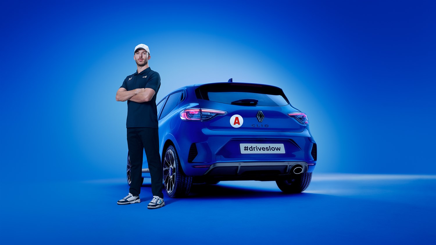 Маж во спортска облека стои до сино  Renault Clio, возилото кое ја носи ознаката А и слоганот  #driveslow.